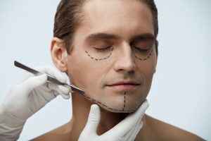 A man with markings on his face ans someone handling a scalpel up to his jaw.